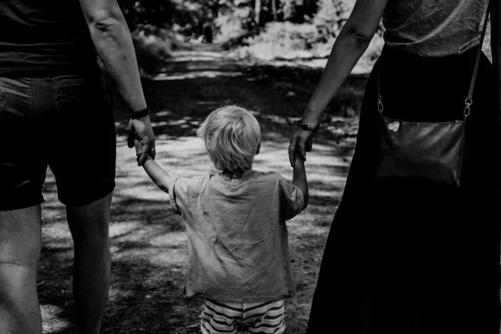 Photographie en noir et blanc montrant un jeune enfant vu de dos, tenant la main de deux adultes de chaque côté, marchant sur un chemin bordé de nature. L’image évoque le lien parental, la protection et ce qui se transforme profondément dans la vie d’un enfant après la mort de ses parents.